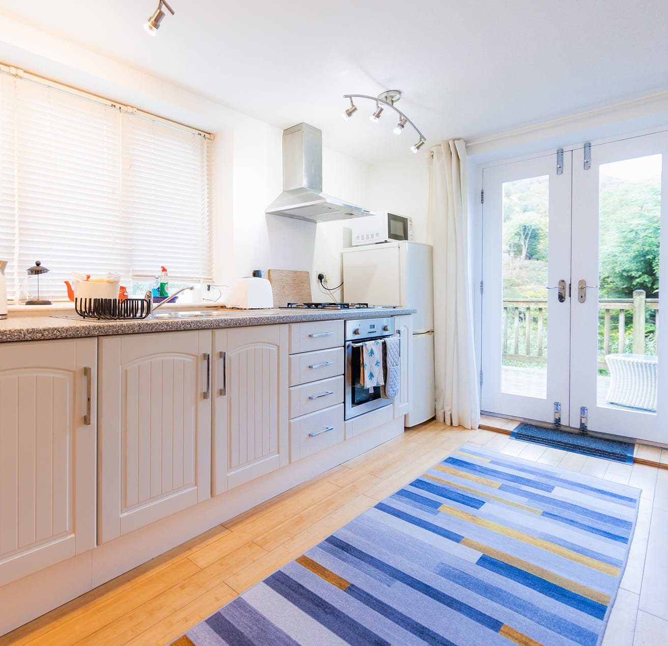A bright kitchen with white cabinets, a granite countertop, and a window with closed blinds. There's a stove, a microwave on top of a refrigerator, and a door leading outside to a deck with greenery. A colorful striped rug is on the wooden floor.