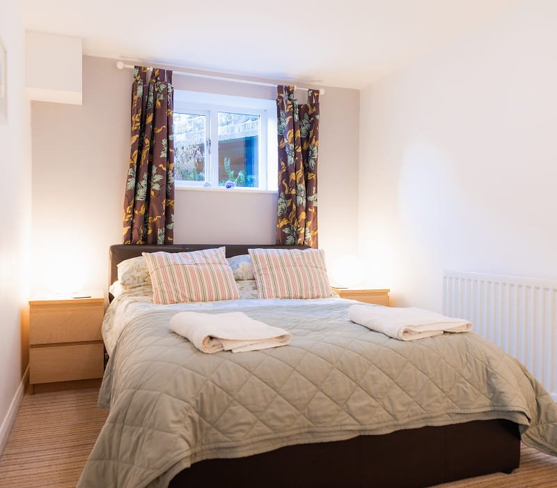 A tidy bedroom with a bed, beige quilt, two striped pillows, two towels, wooden bedside tables, a window with floral curtains, and white walls.