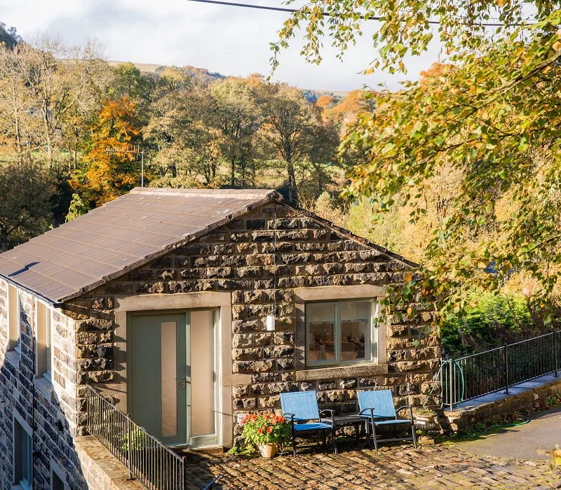 Small stone house with a sloped roof, two windows, a white door, outdoor chairs, and trees with autumn foliage in the background.