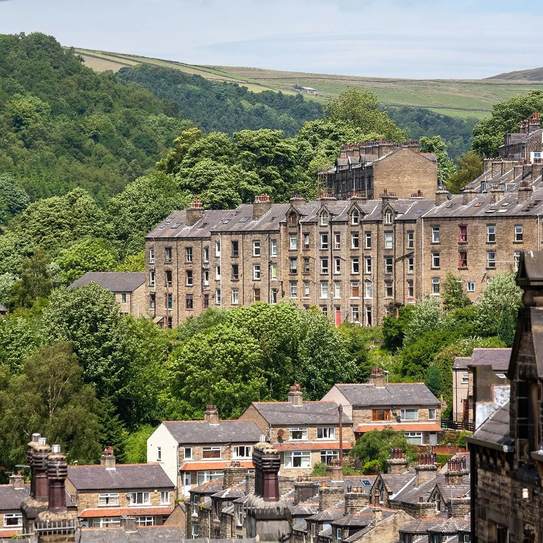 A scenic view of a hillside town with numerous old brick buildings and houses surrounded by lush green trees and rolling hills in the background.