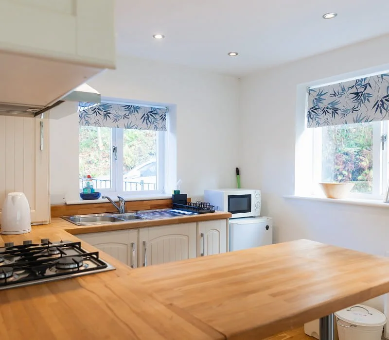 Bright kitchen with wooden counters, white cabinets, a stove, a microwave on a small refrigerator, and two windows with blue and white leaf-patterned blinds.