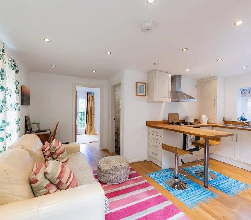 Open-concept living room and kitchen with a beige sofa, striped pillows, colorful rugs, and wooden furniture.
