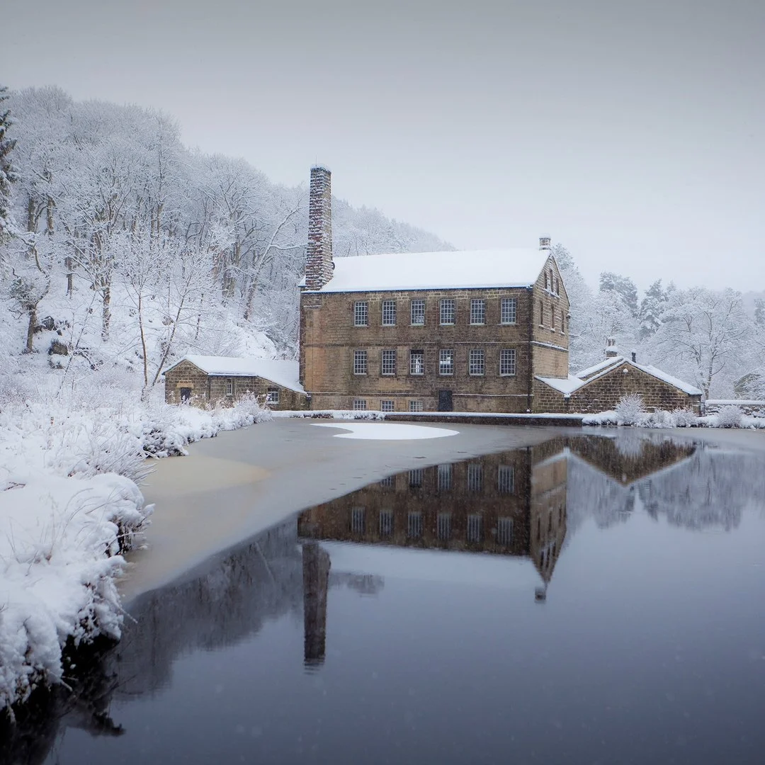 A large stone house surrounded by snow-covered trees with a chimney, beside a partially frozen pond that reflects the house and trees.