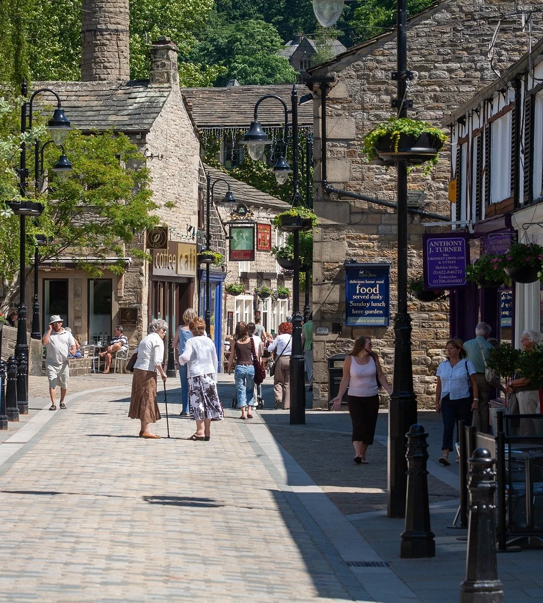 A bustling cobblestone street lined with shops and cafes, with people walking and interacting outdoors on a sunny day. Traditional stone buildings with hanging flower baskets and street lamps add to the charming atmosphere.