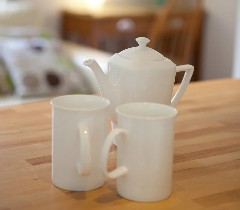 Two white ceramic mugs and a white ceramic teapot on a wooden table in a cozy kitchen.