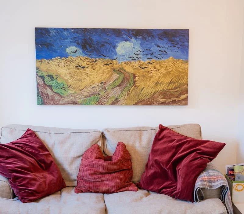 A living room with a beige sofa decorated with three red pillows and a 'paw print' shaped pillow. Above the sofa, there is a large painting of a landscape with wheat fields, a dirt path, and a sky with clouds and birds.