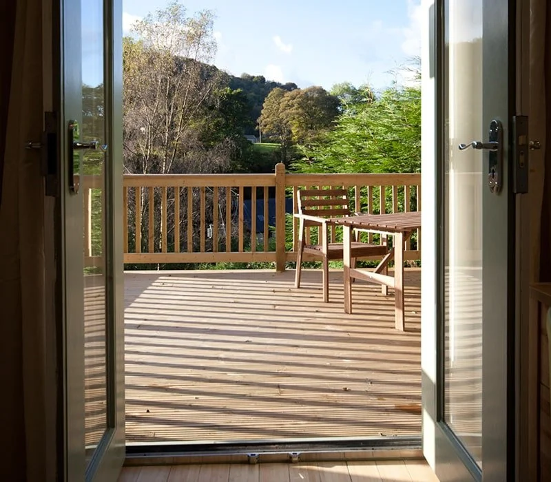 View of a wooden deck with a chair and railing, looking out to trees and hills under a partly cloudy sky, seen from inside a house through open glass doors.