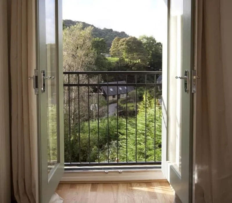 Open French doors leading to a small balcony overlooking a lush green landscape with trees, hills, and a few rooftops.