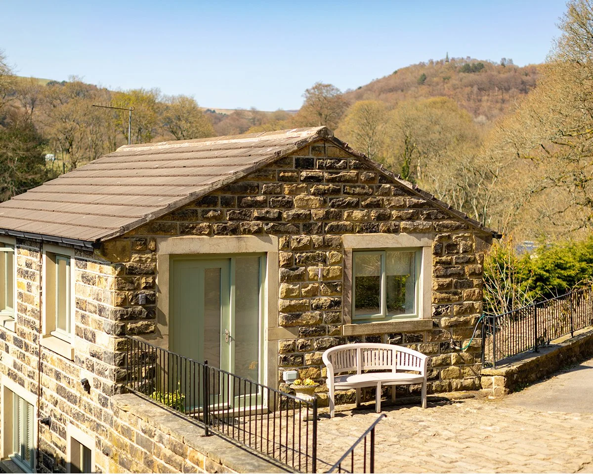 Stone house with slate roof and a small balcony with black railing, situated in a scenic area with trees and hills in the background.