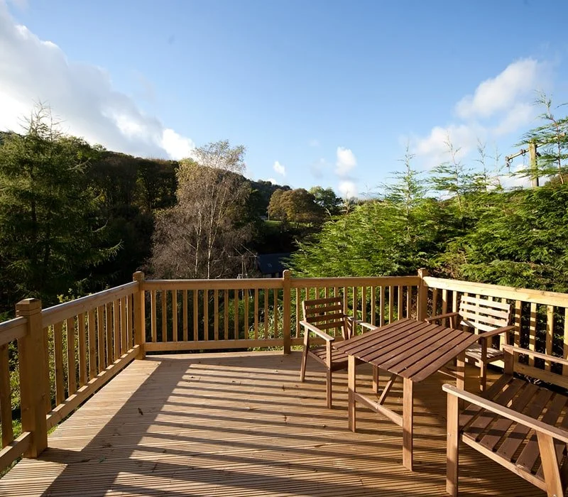 Wooden deck with four wooden chairs and a wooden table, overlooking a scenic view of trees and a hilly landscape under a partly cloudy sky.