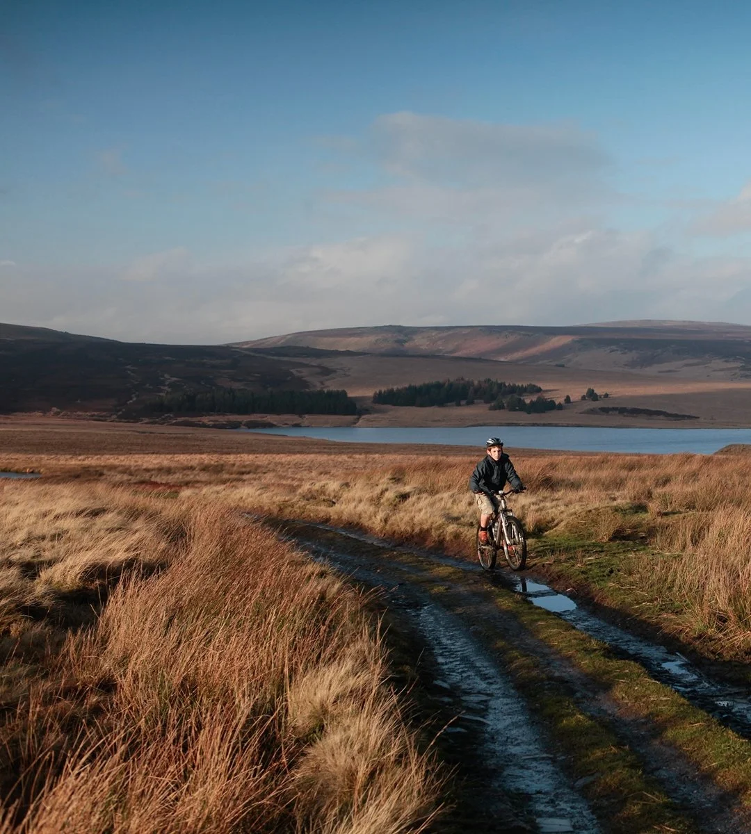 A person riding a mountain bike on a muddy trail through tall grass in a rural landscape with a lake and hills in the background.