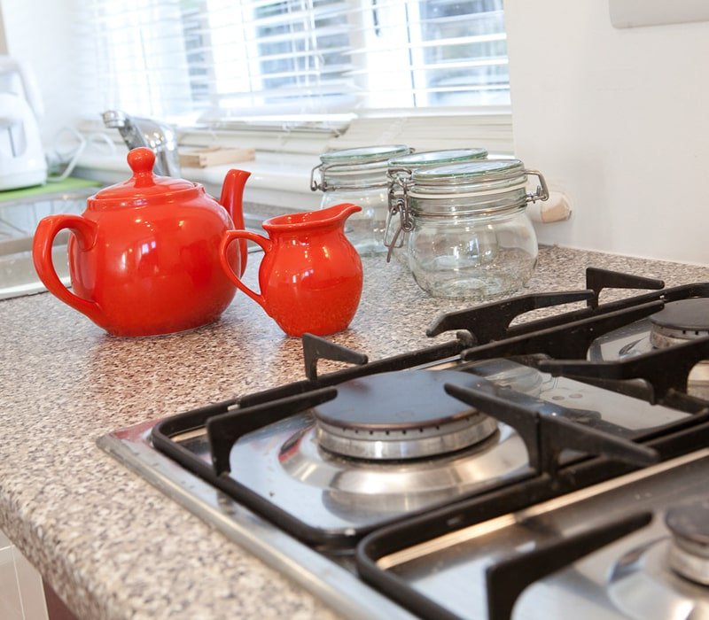 Red teapot and sugar bowl on kitchen countertop with jars and a gas stove in the background.