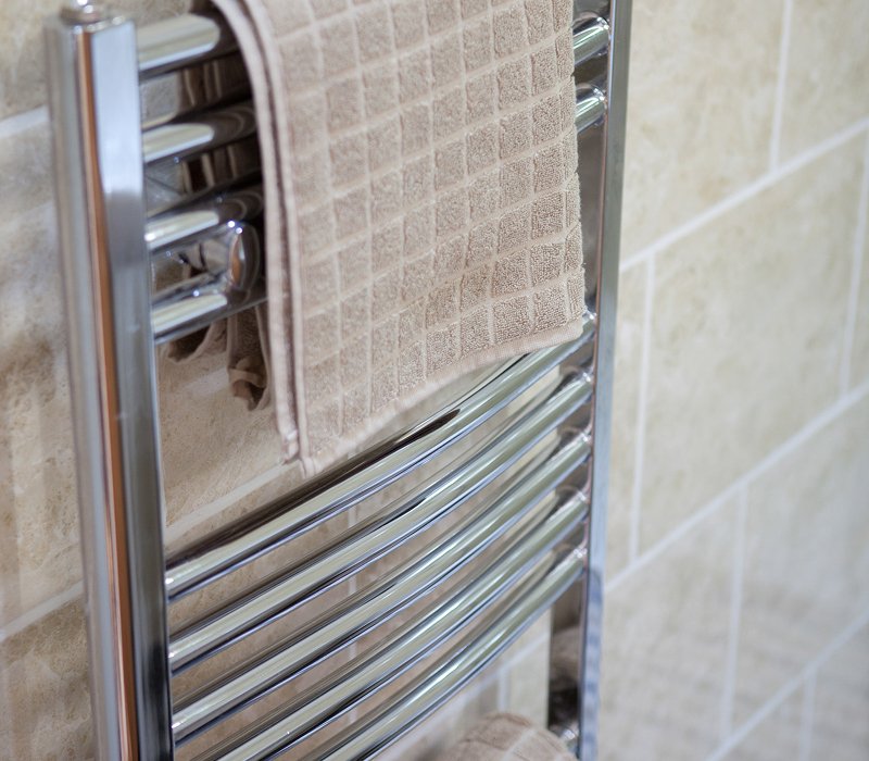 Close-up of a stainless steel towel radiator with a beige towel hanging on it, mounted on a beige tiled bathroom wall.
