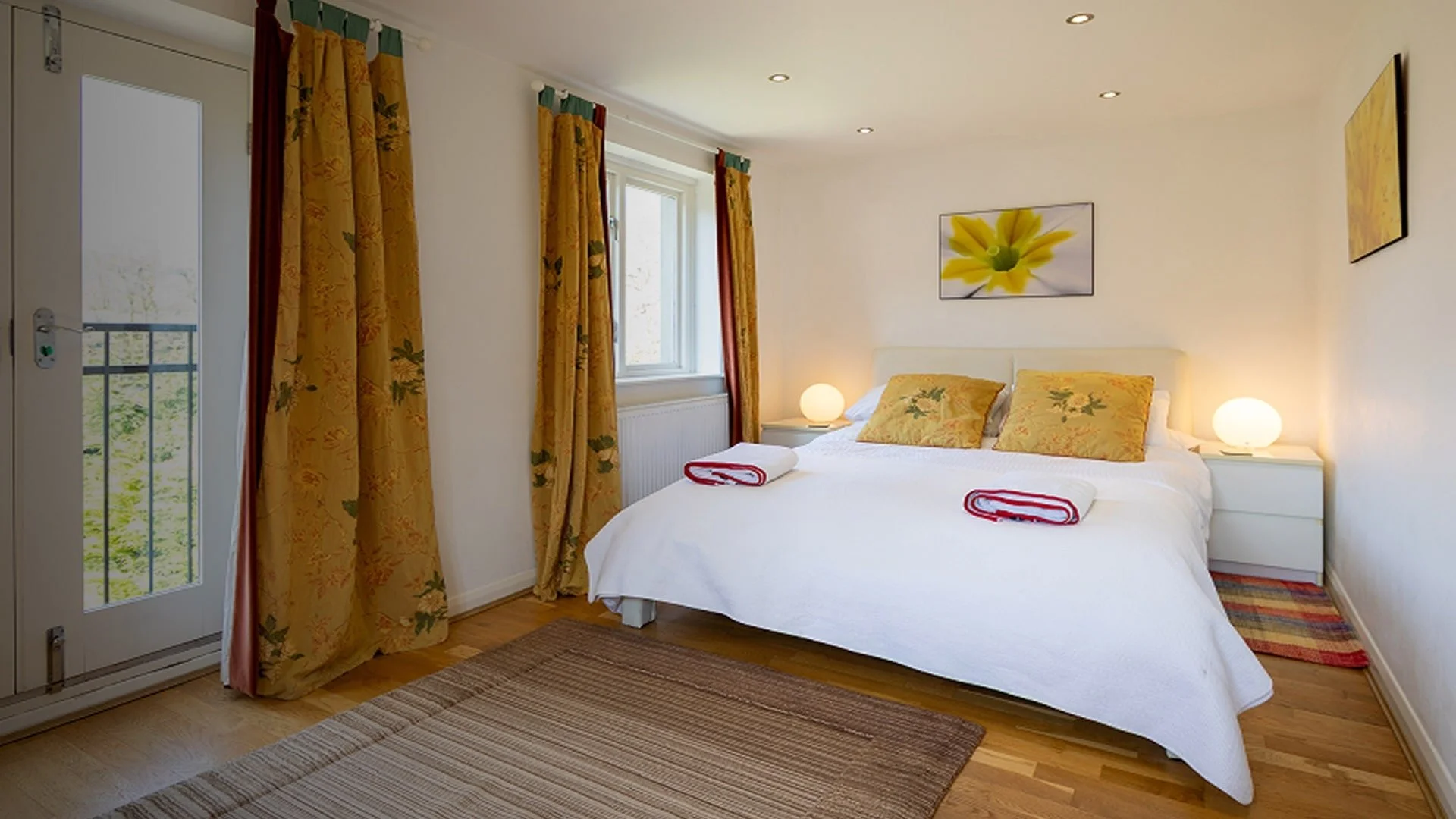 A cozy bedroom with a white bed, yellow floral pillows, and two rolled towels on the bed. There are two bedside tables with glowing lamps, a window with yellow floral curtains, and wall art featuring a yellow flower. The room has wooden flooring and a light, airy atmosphere.