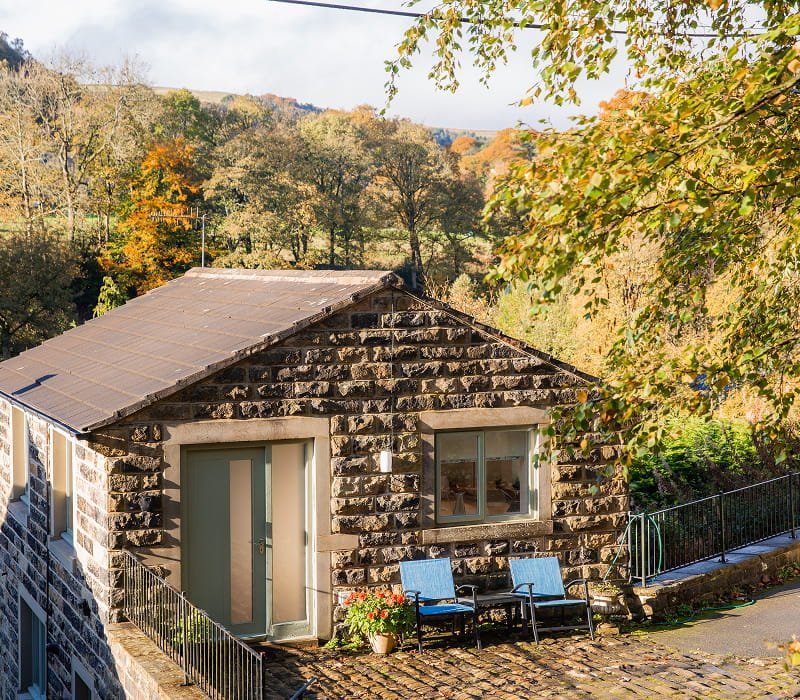 Stone house with a sloped roof, two windows, a potted plant, and two blue chairs outside on a brick patio surrounded by trees with fall foliage.