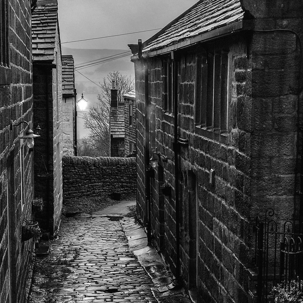 A narrow cobblestone alleyway between old stone buildings on a foggy evening, illuminated by a street lamp.