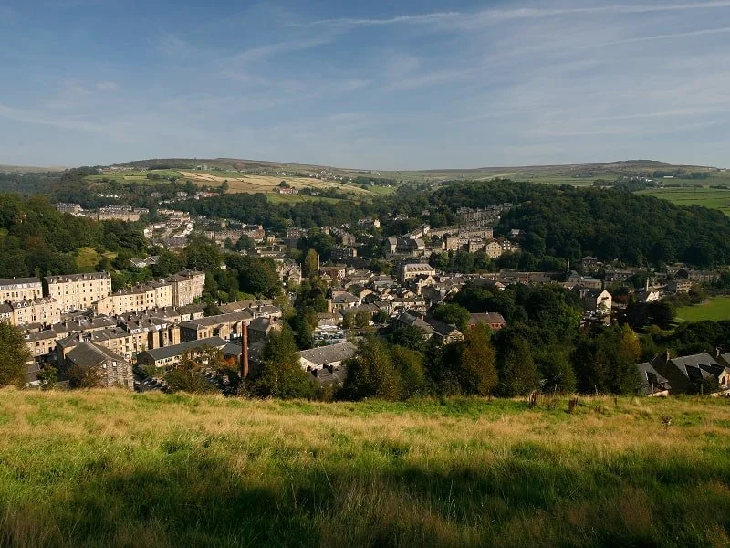 A scenic view of a small town surrounded by green hills, with houses and buildings nestled among trees under a partly cloudy sky.