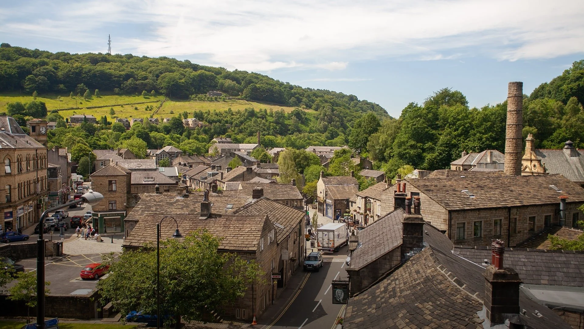 View of a small town with stone buildings and rooftops, surrounded by green hills and trees