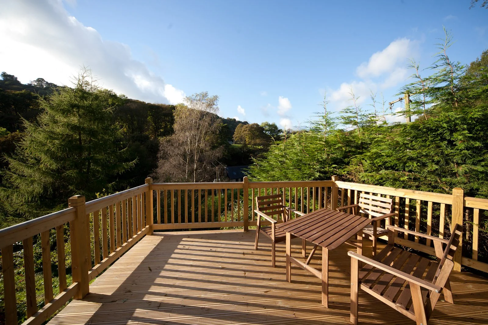 Wooden deck with outdoor furniture overlooking a lush green landscape with trees and a blue sky.