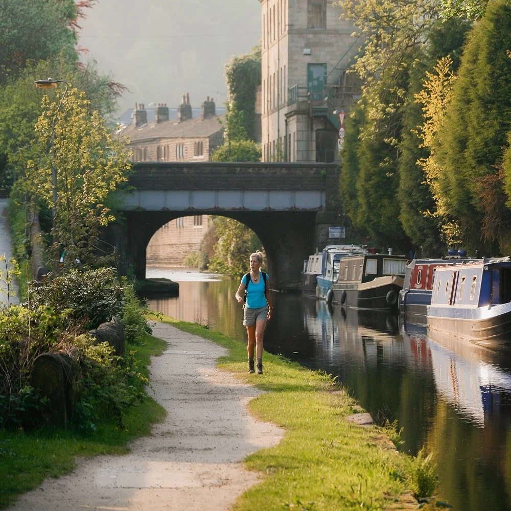Woman walking along a canal path with boats on the water, trees on one side, buildings in the background, and a bridge overhead in the late afternoon or early evening.