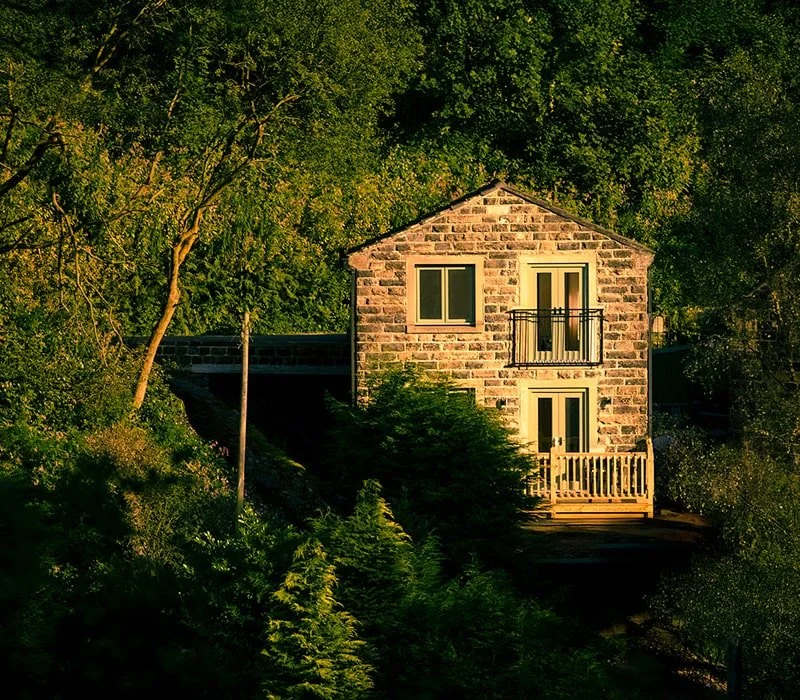 A two-story house made of brick and stone, surrounded by green trees and foliage, with a small balcony on the second floor and a porch on the first floor.