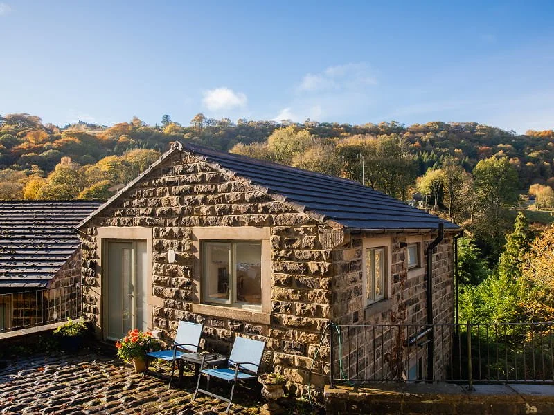 Stone cottage with a dark roof and outdoor seating, set against a backdrop of rolling hills and trees with autumn foliage.