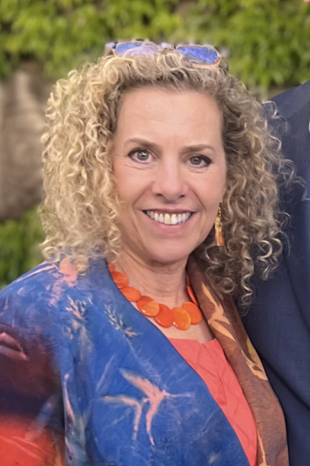 A woman with blonde curly hair wearing sunglasses on her head, orange necklace, and a colorful patterned blazer, smiling outdoors.
