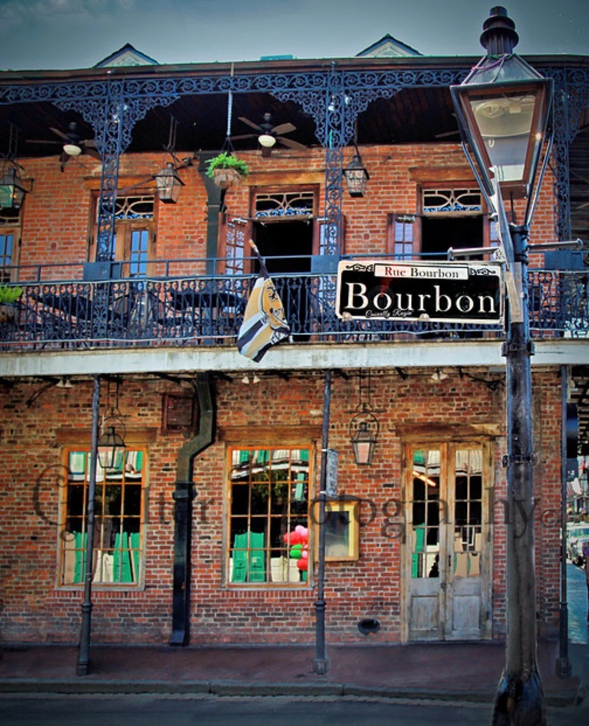 Brick building with two floors, black wrought iron balcony and ornate balcony railing, hanging lamps, colorful balloons, street signs for Rue Bourbon, and a vintage-style street lamp in the foreground.