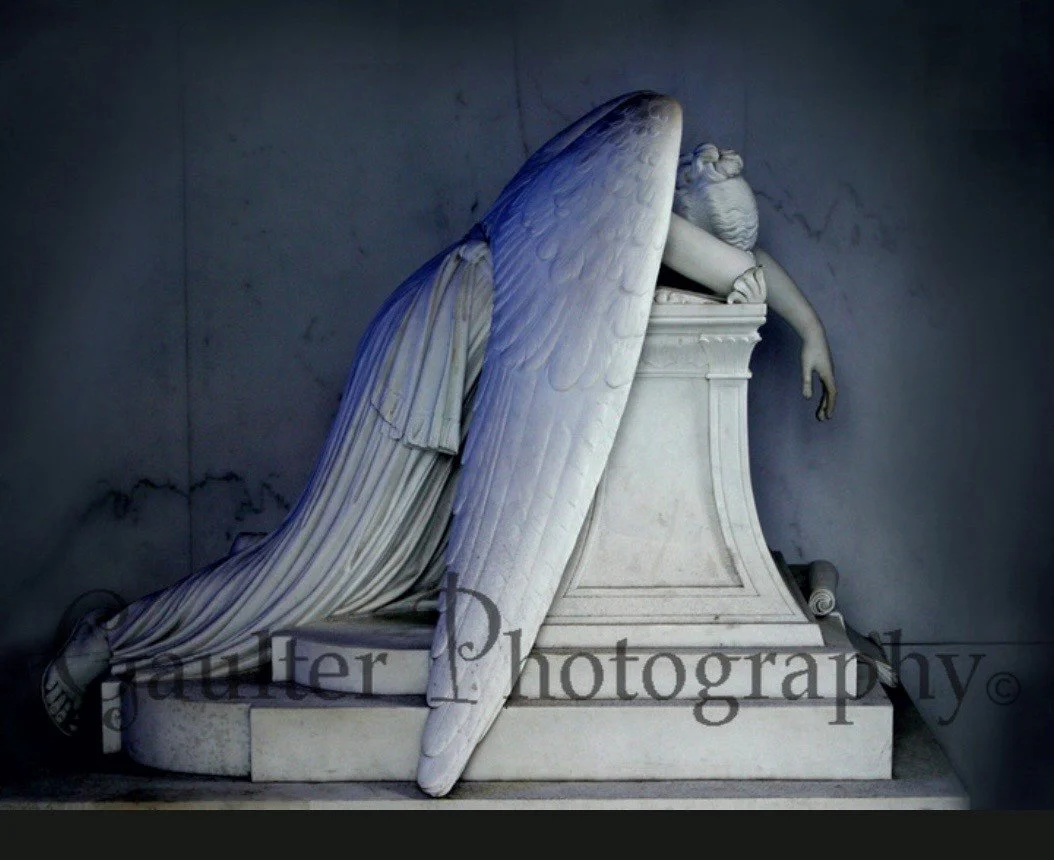 Marble statue of a fallen angel with wings, resting its head and arm on a pedestal.
