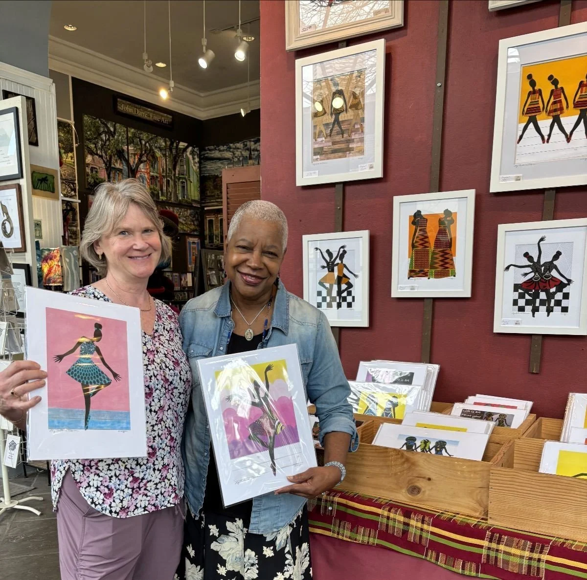 Two women standing in an art gallery, holding framed artwork,  other art displayed behind.