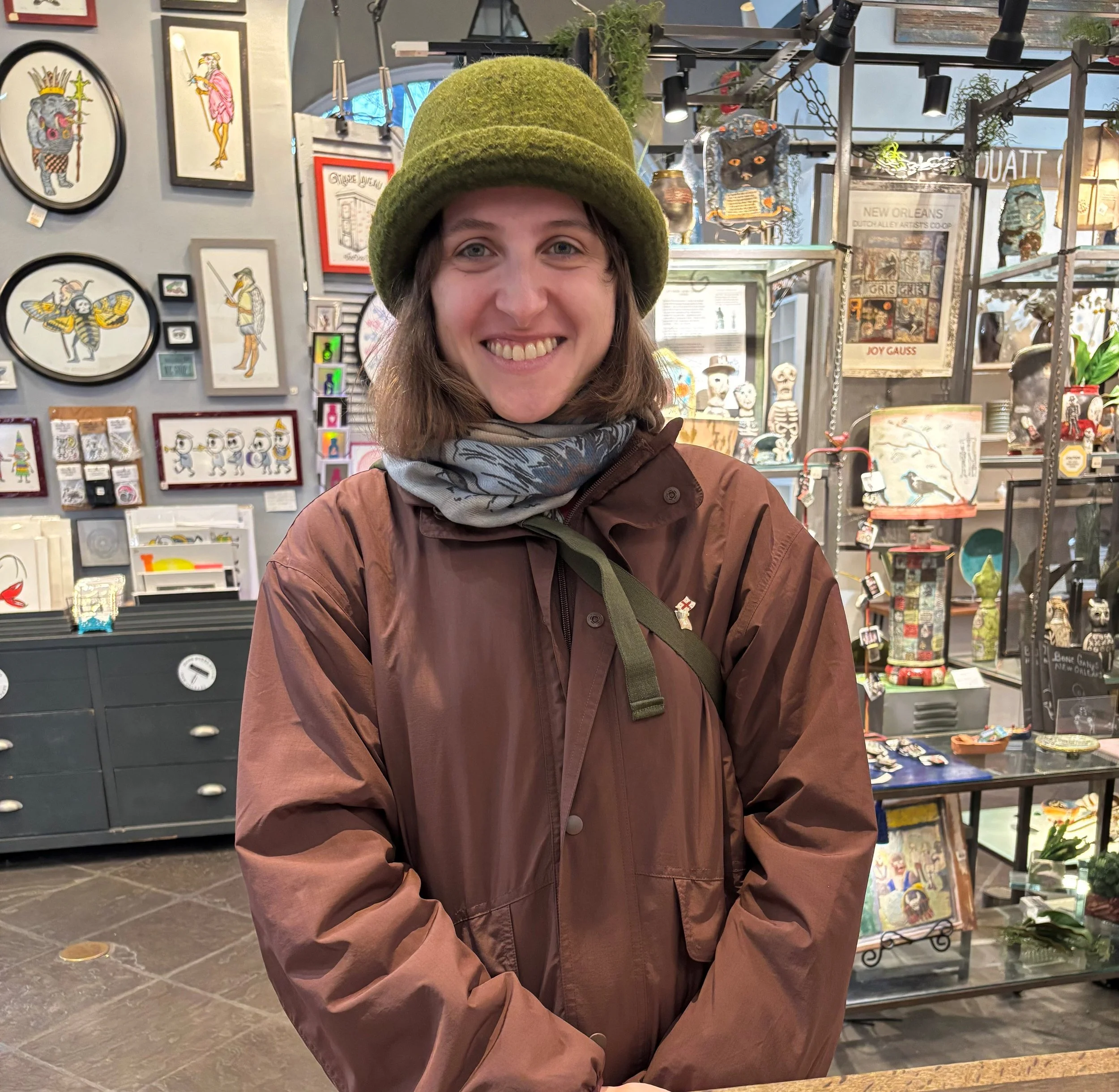 A woman wearing a green hat and brown jacket smiling inside an art shop with framed artwork, sculptures, and decorative items displayed on the walls and shelves.