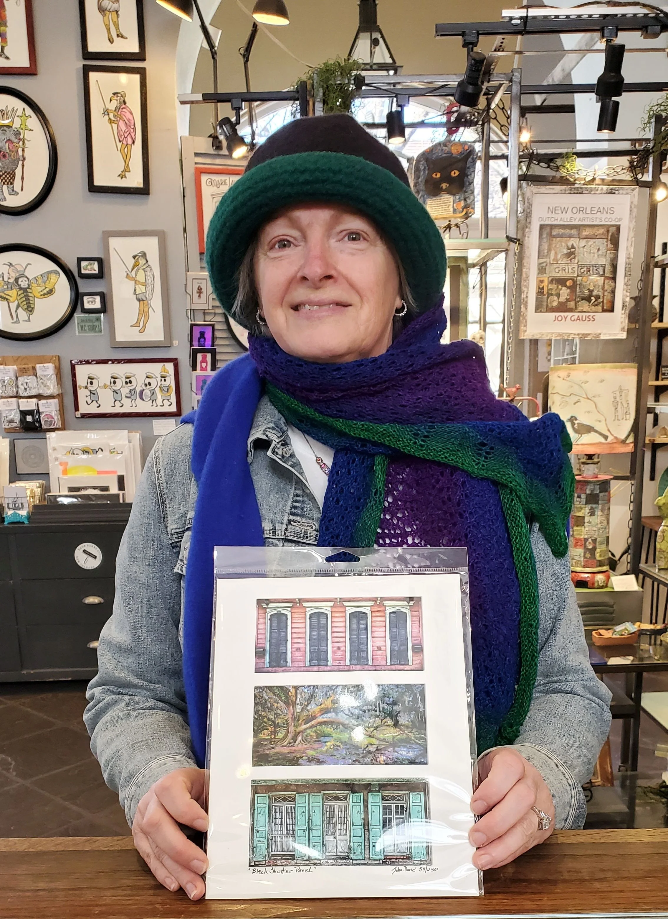 A woman wearing a green and purple knitted scarf and a denim jacket holding a framed artwork of a blue and red building with large windows. The background shows art on the walls, including oval and rectangular framed illustrations of animals and people, and shops with various items.