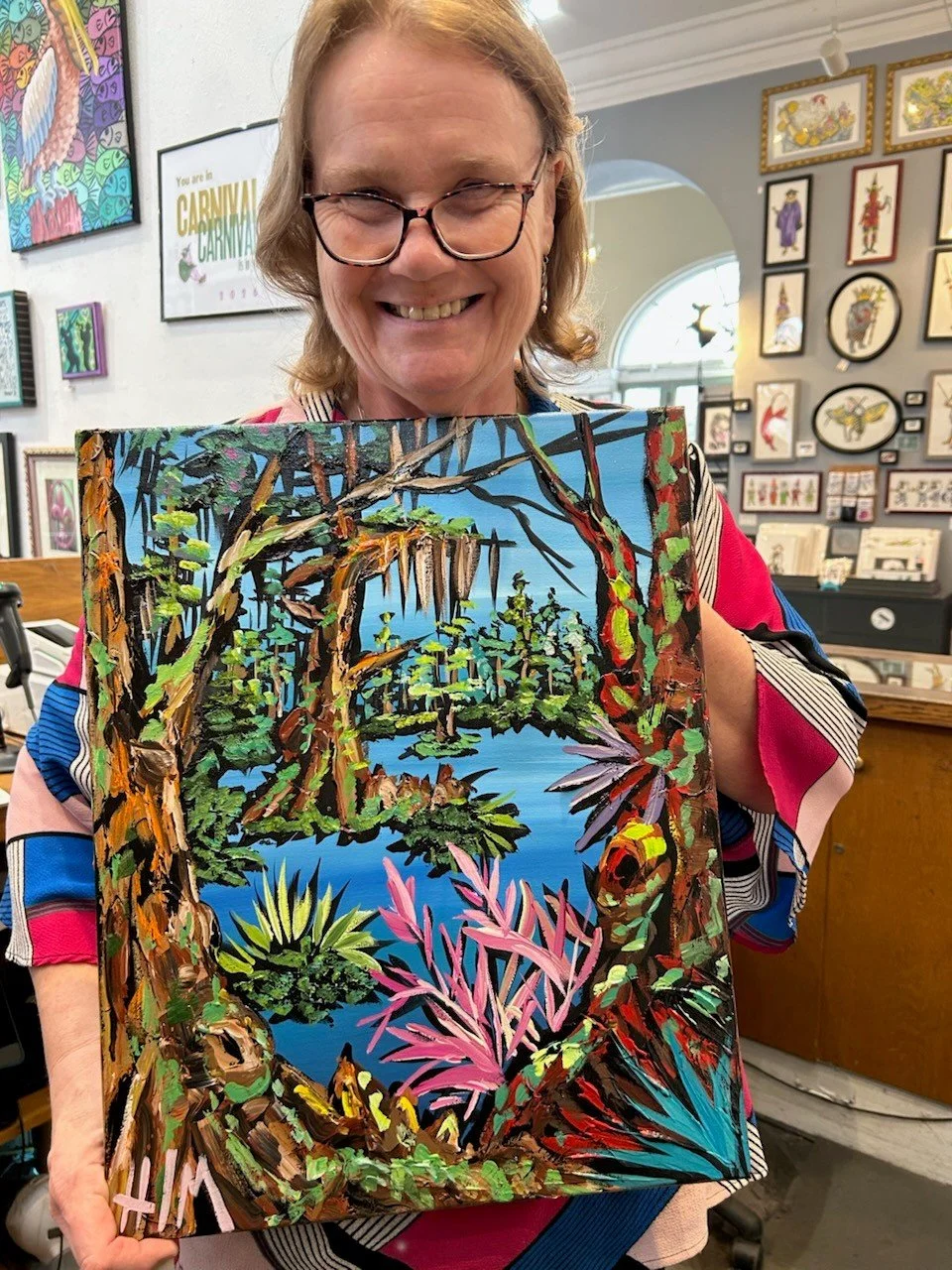 Woman wearing glasses and colorful clothing holding a vibrant painting of a jungle scene with trees, green foliage, and a body of water, inside an art gallery filled with various framed artworks on the walls.