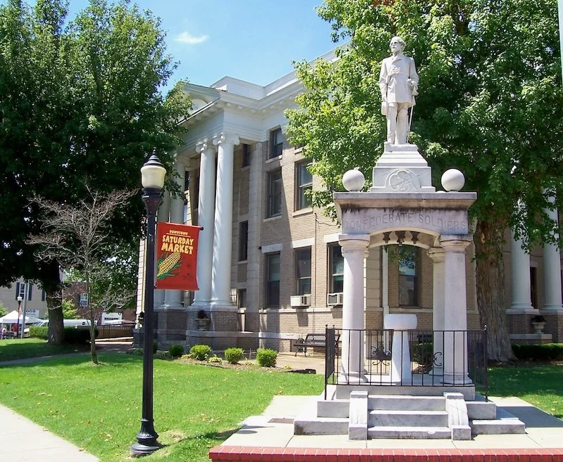 Statue of a man in military uniform on a pedestal with an inscription 'Union Defeated Soldiers.' The statue is situated in front of a large white building with columns, surrounded by green trees and grass, with a lamp post nearby.