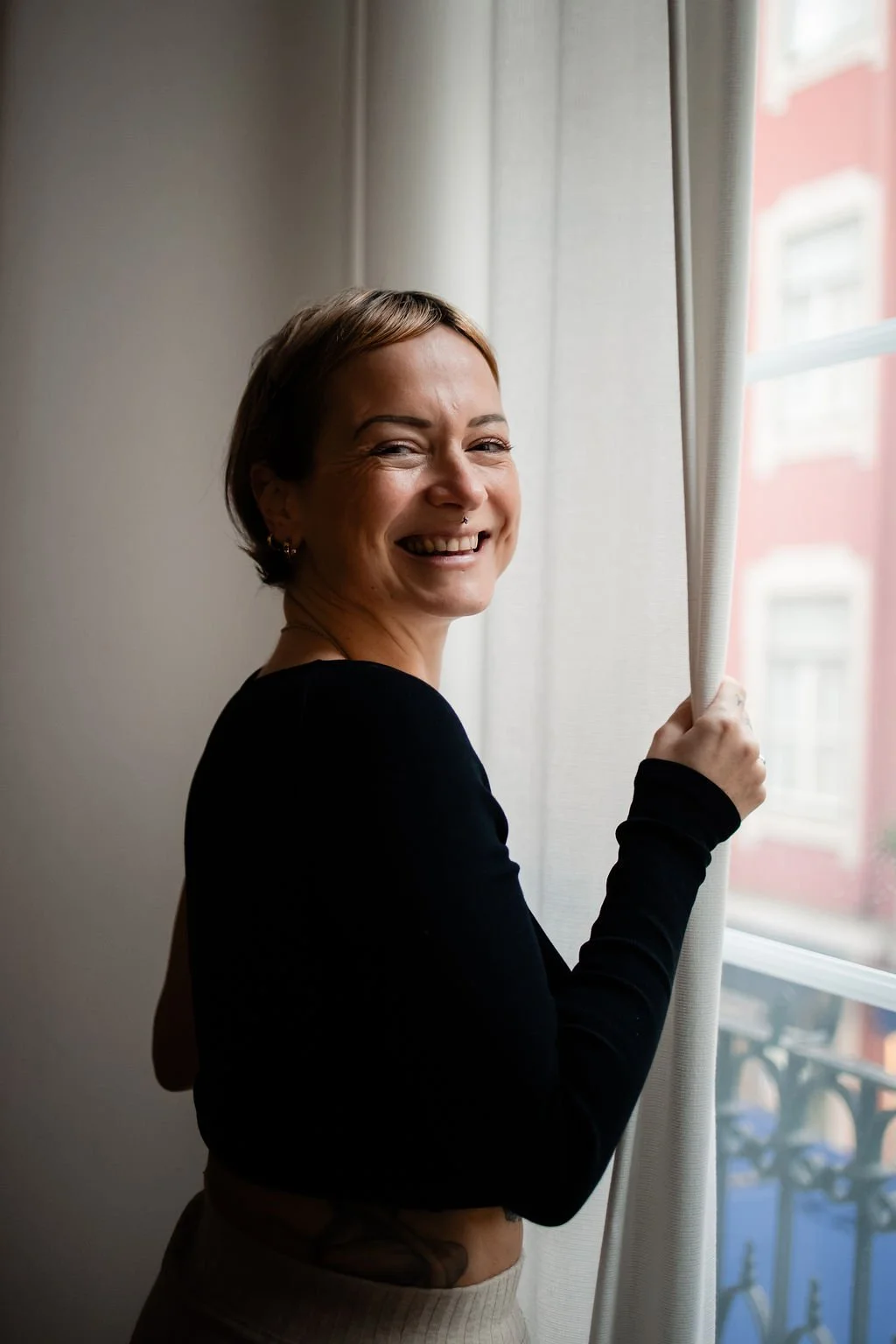 Smiling woman with short hair looking at camera while holding a curtain near a window.