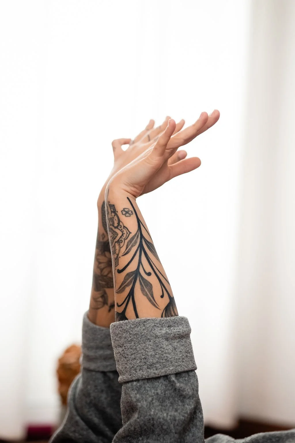 Person with tattoos on forearm, gray long-sleeve shirt, raised hand with fingers spread against a plain white background.