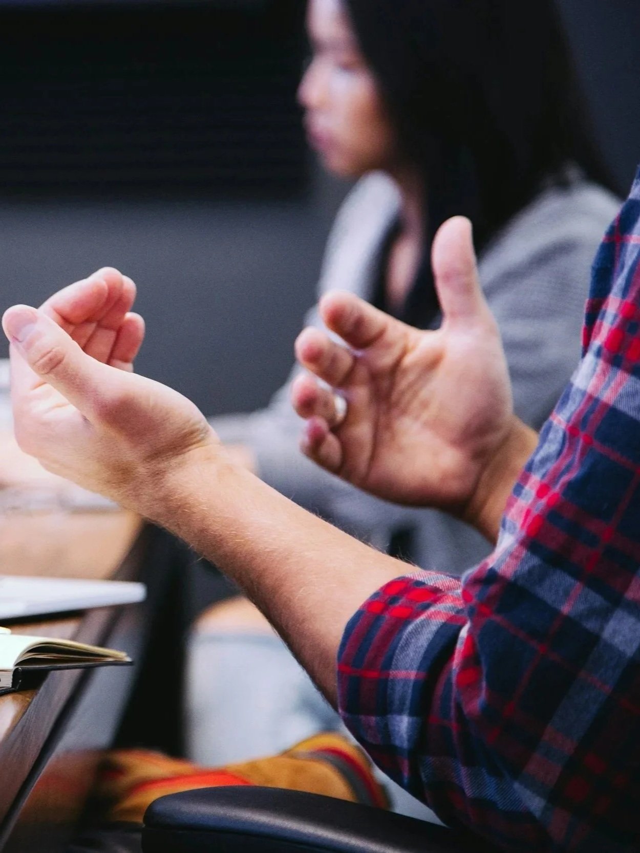 Nahaufnahme von zwei Menschen, die miteinander sprechen, mit Fokus auf ihre Hände und eine Person im Hintergrund, die auf einem Tisch sitzt.