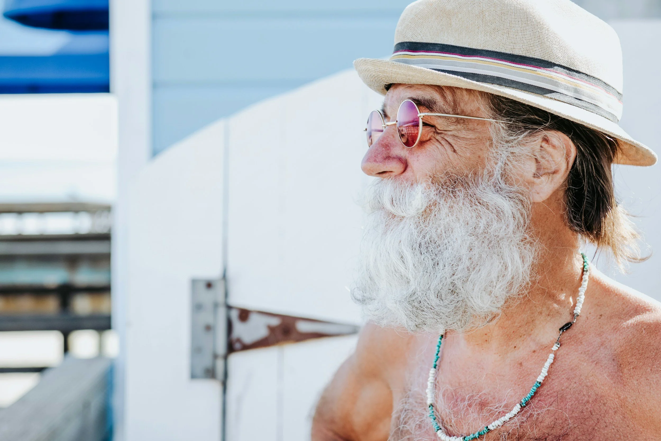 An elegant older man with white beard and mustache, wearing round sunglasses, a light-colored fedora hat, and a beaded necklace.