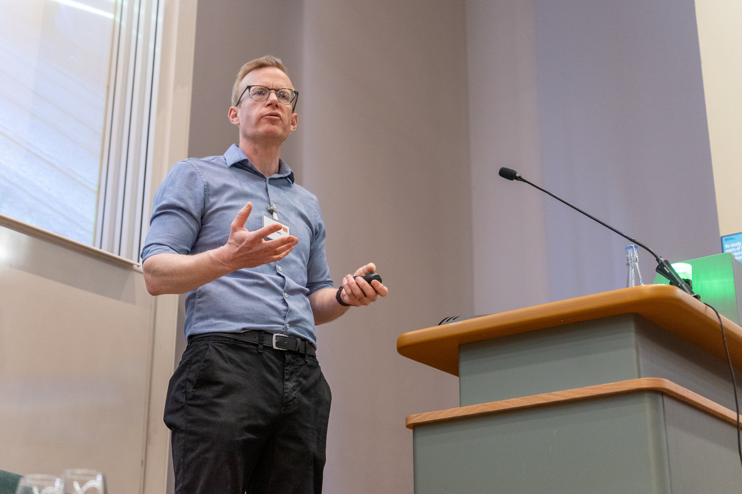Dr Kenton Lewis MBE presentation in a conference room. He is stodd next to a lectern with a microphone.