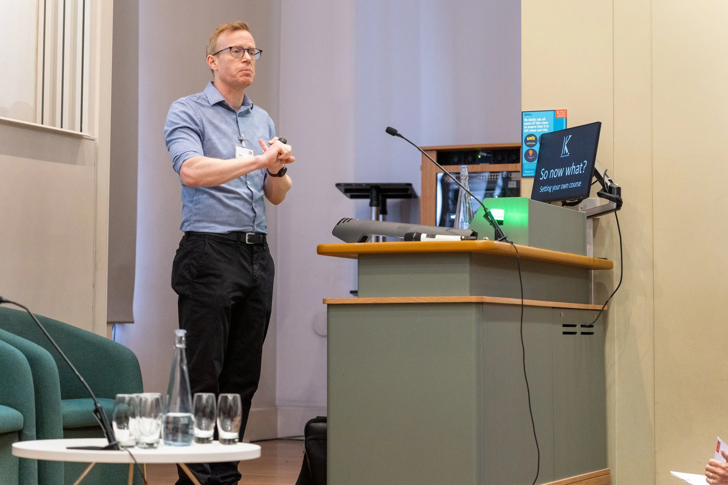 Dr Kenton Lewis MBE presenting at a conference. He stands next to a podium with a monitor displaying the slide titled 'So now what? Setting your own course.'