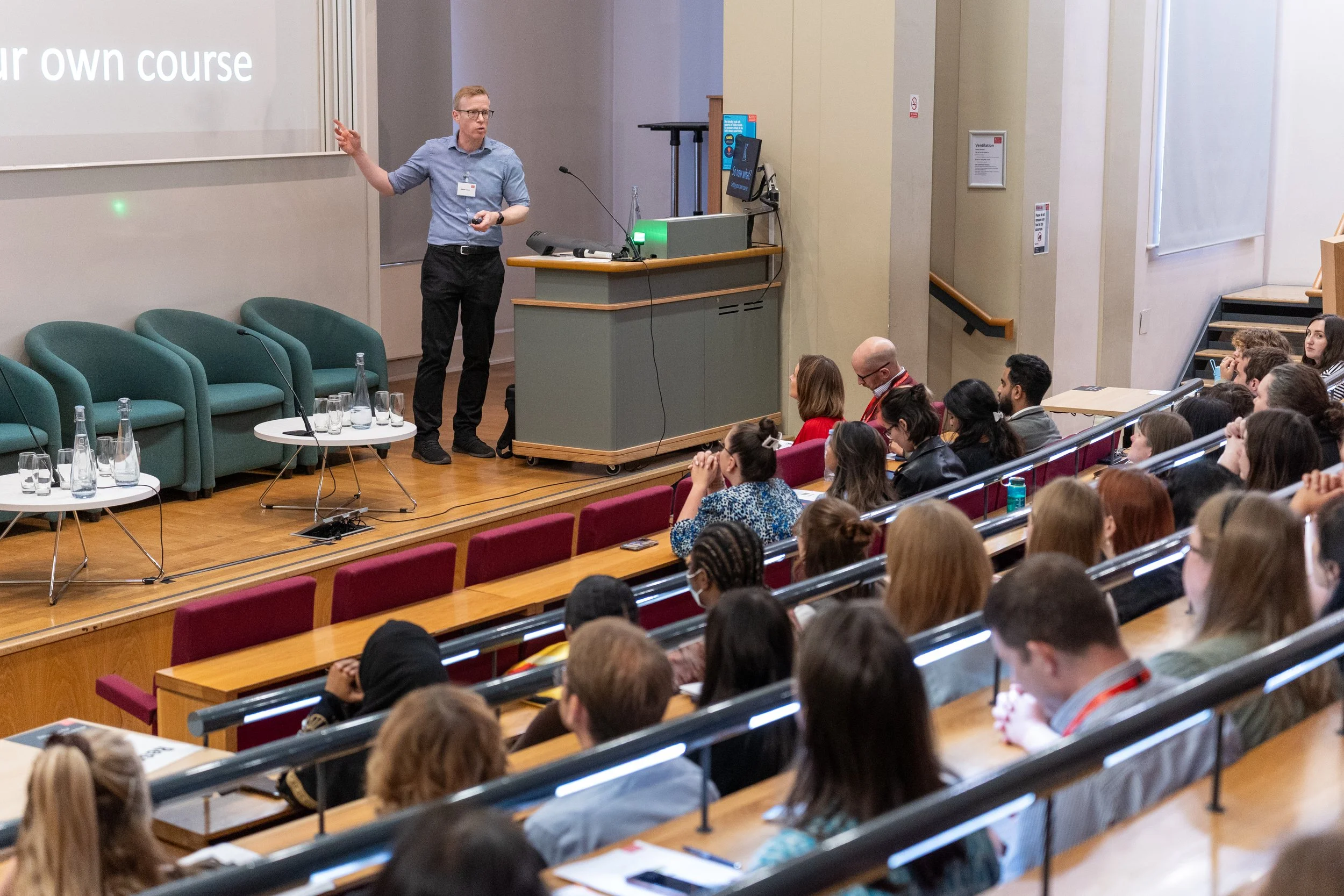 Dr Kenton Lewis MBE presenting to a large audience in a lecture hall.