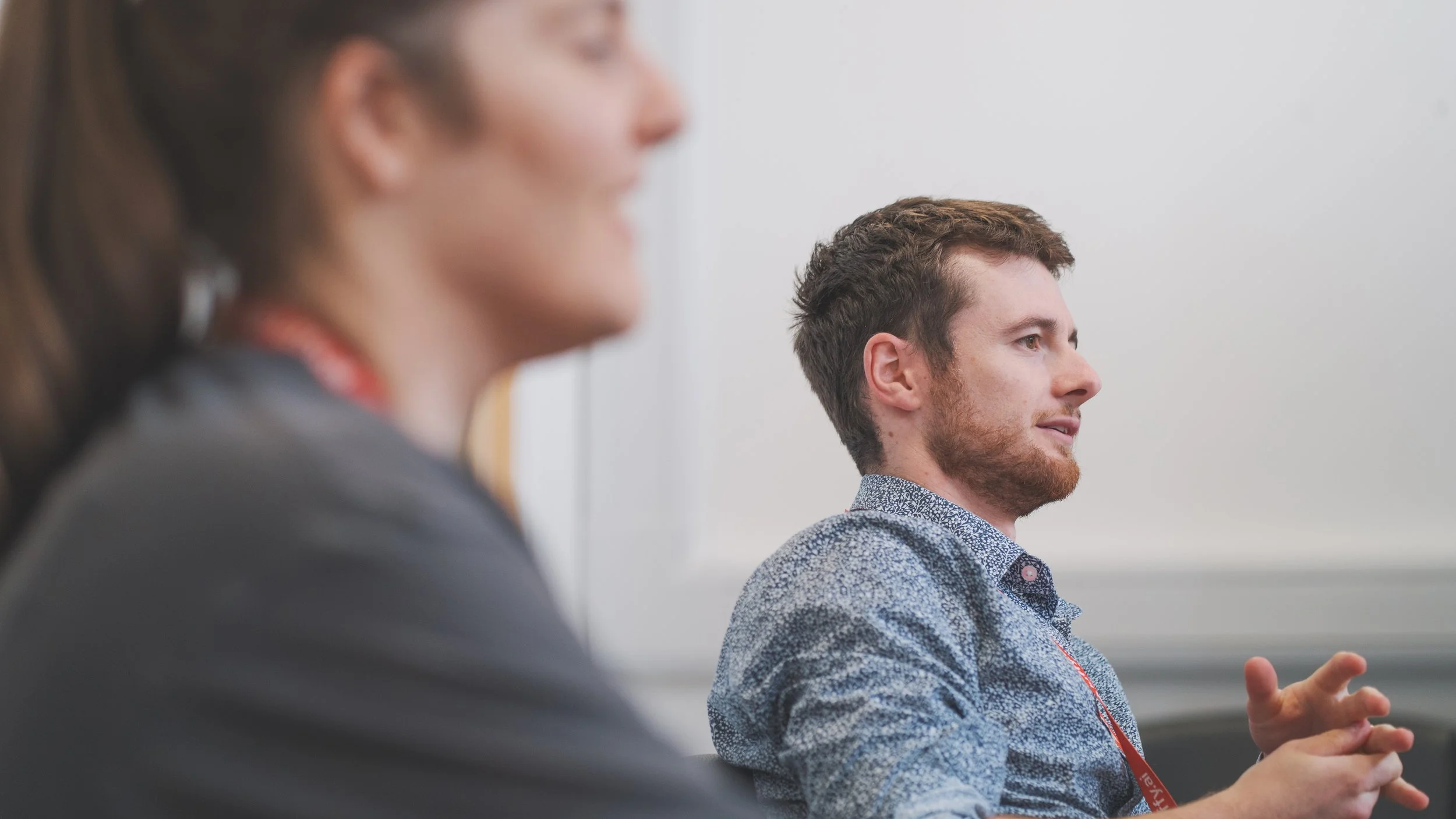 A person with brown hair and a beard, wearing a patterned shirt, is sitting in a meeting room and speaking. A person with brown hair pulled back, is sitting in the foreground, slightly blurred.