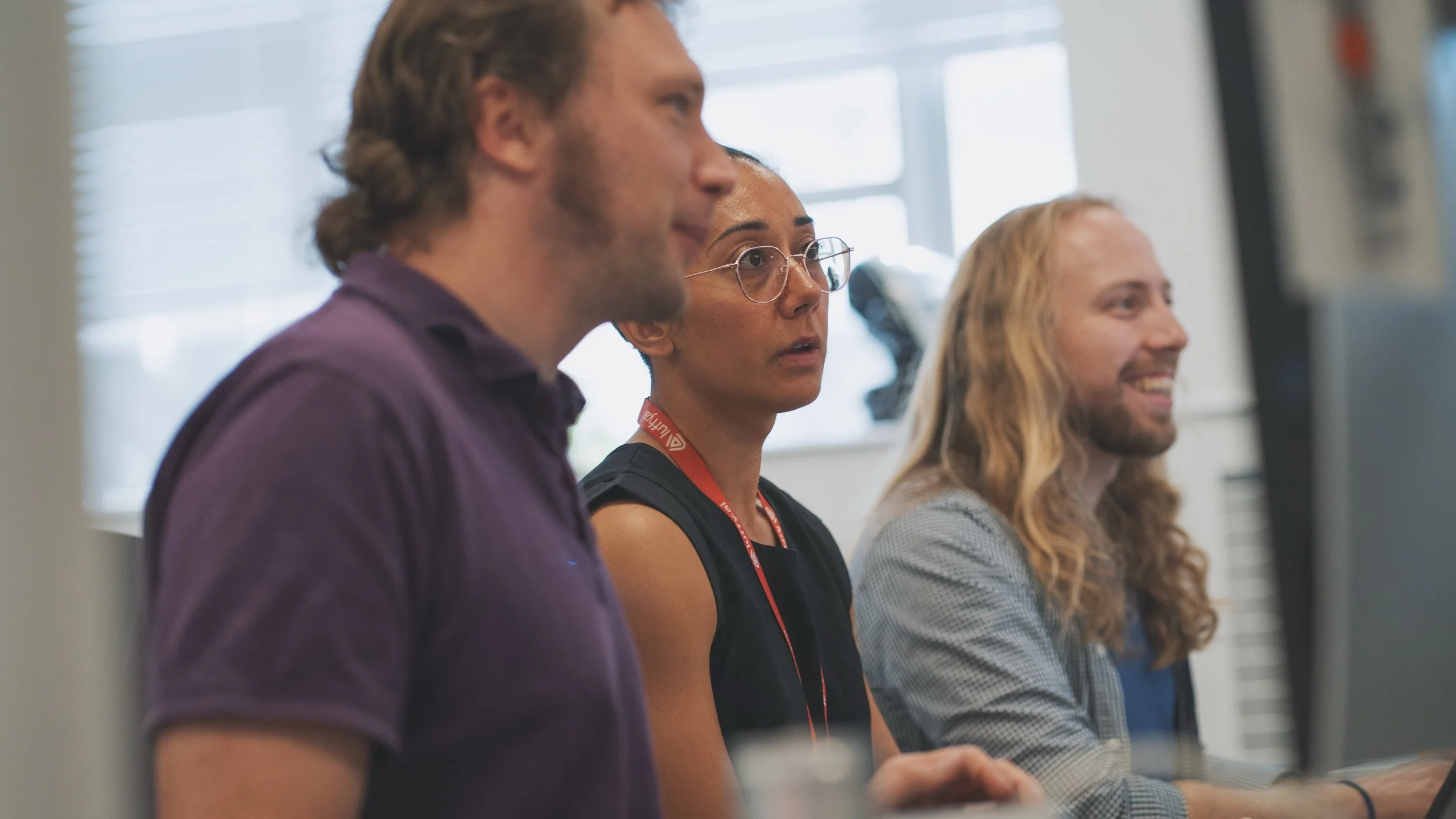 Three people sitting at desks in front of computer monitors, engaged in work or discussion in an office setting.