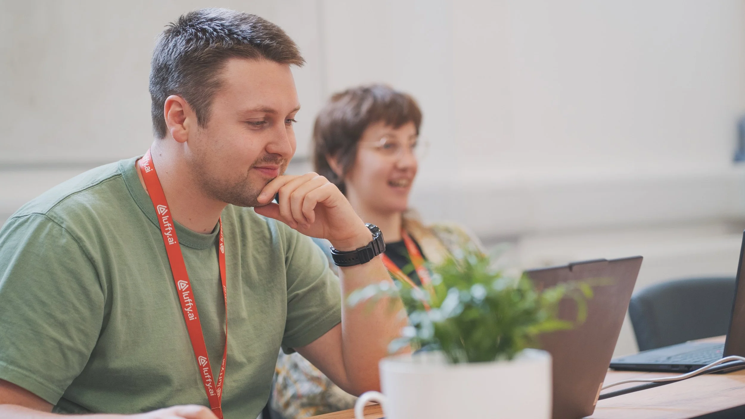 Two people sitting at a table in an office, using laptops, with a potted plant in the foreground. 