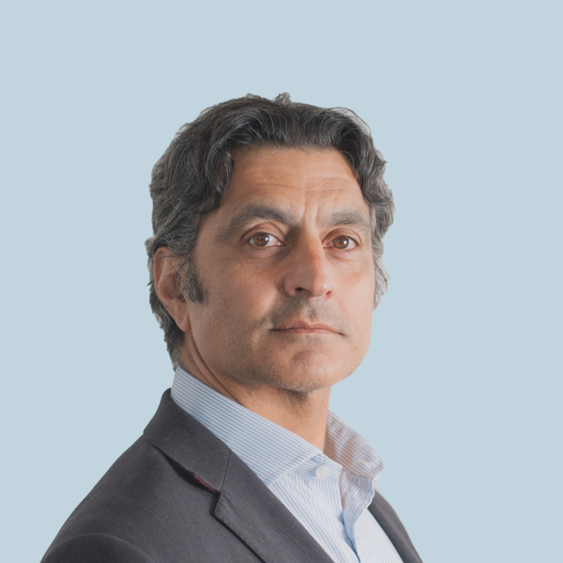 Professional man with wavy dark hair wearing a dark suit and light blue collared shirt, looking serious, against a light blue background.