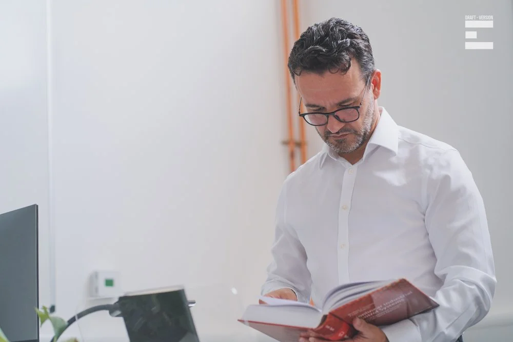 A man with glasses and dark hair reading a book in an office.