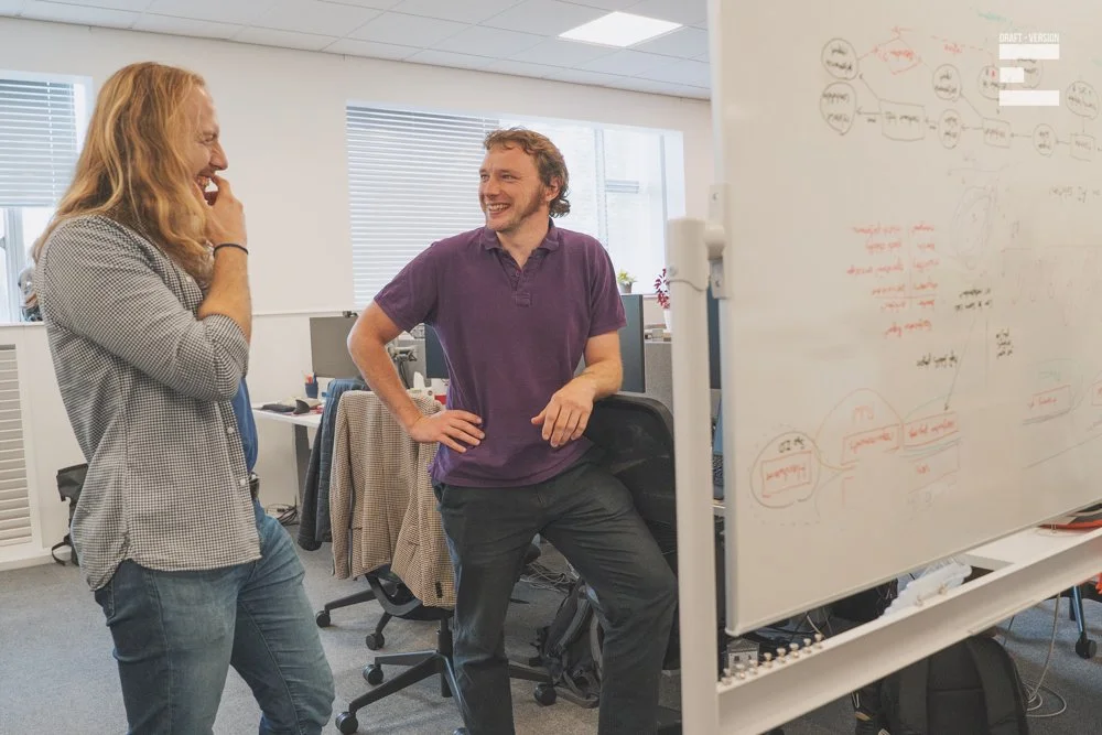 Two colleagues, laughing and talking in an office, standing near a whiteboard with diagrams and notes.