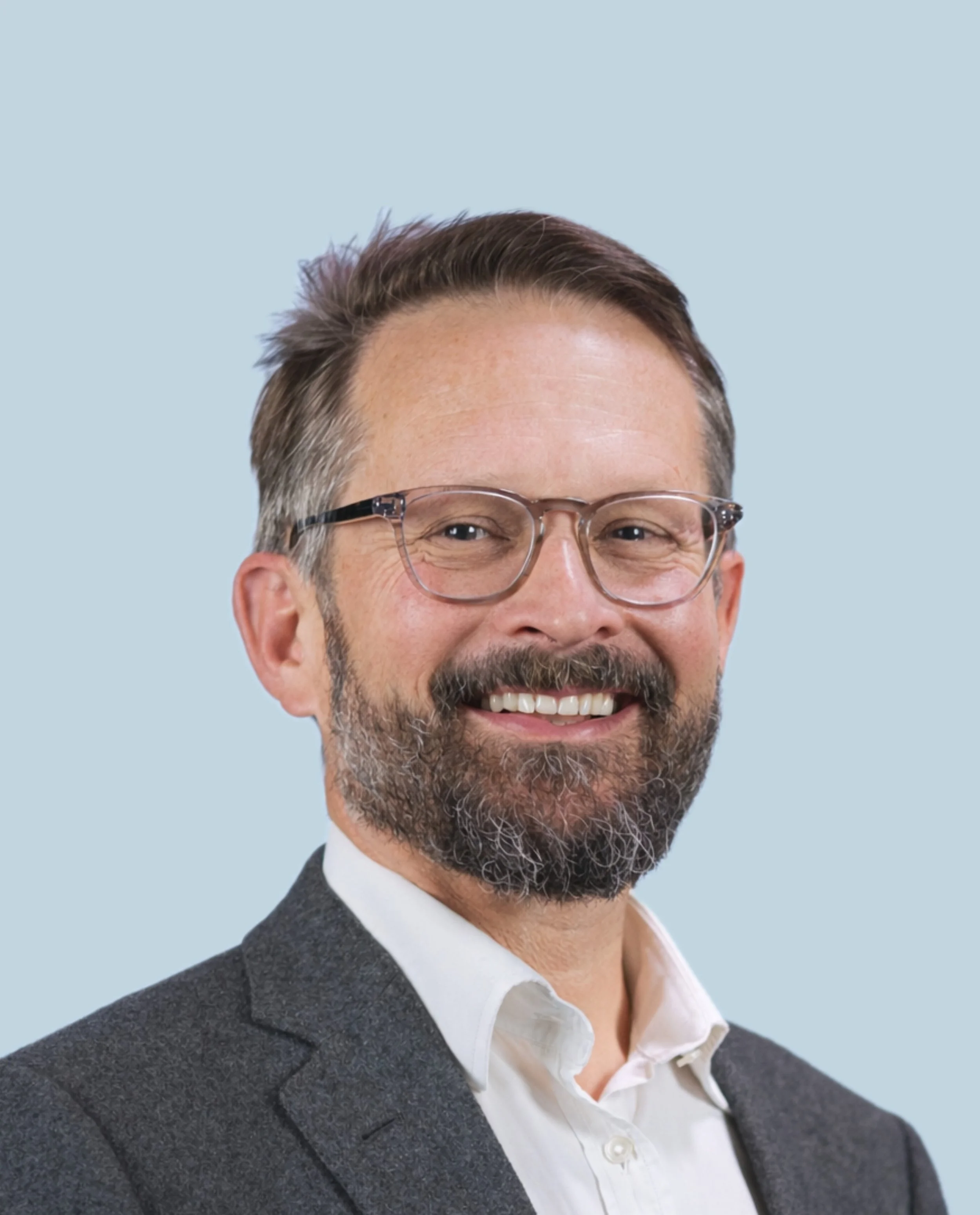 A smiling man with glasses, a beard, and a suit against a light blue background.