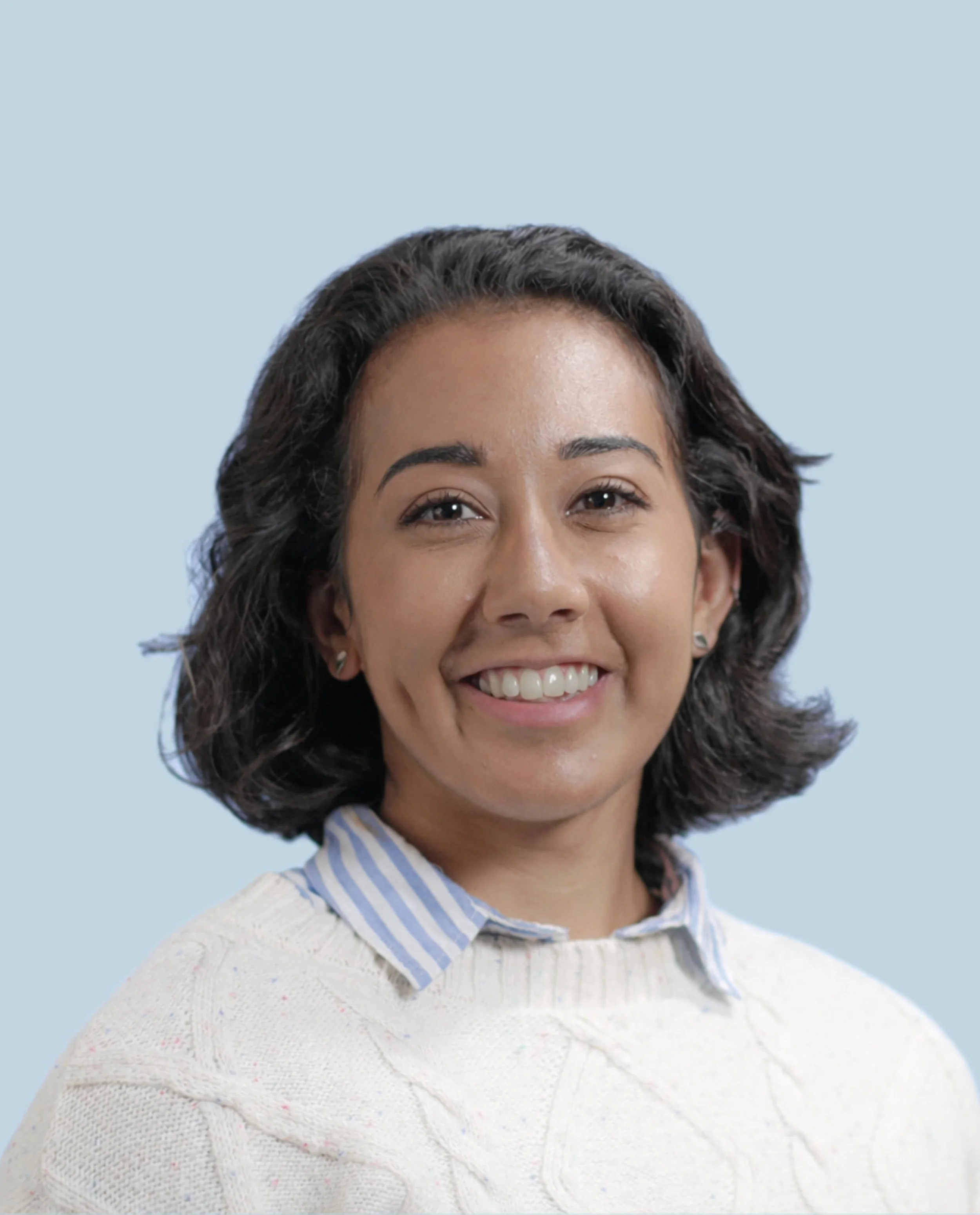Portrait of a woman with dark wavy hair, smiling, wearing a light-colored sweater and a blue striped collared shirt underneath, against a light blue background.