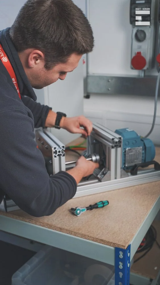 A man working on assembling or repairing a mechanical or electronic device at a workshop table, with tools and machinery.